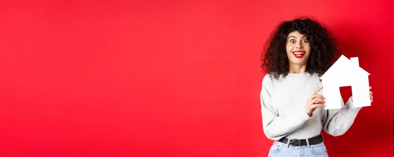 A smiling, excited woman in real estate showing a paper house cutout and looking amazed, standing against a red background.